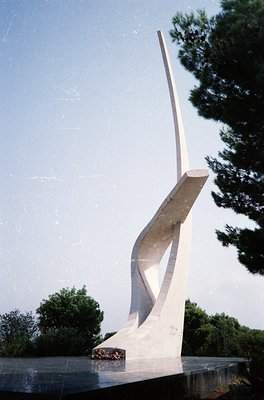 Modernist concrete monument resembling a stylized flame or abstract wing, set on a circular base. Minimalist design with clea...