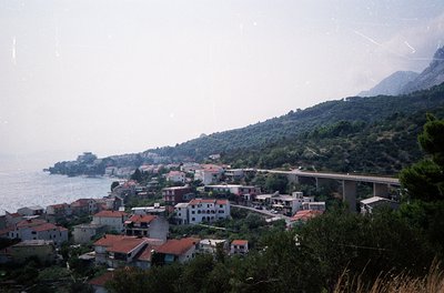 Coastal village nestled between sea and mountains, featuring clustered Mediterranean-style buildings with terracotta roofs. A...