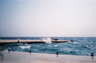 Vintage seaside pool with concrete edge and wooden deck extending into choppy waves. Two figures in swimwear stand near the w...