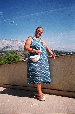 A woman in a 1990s-style polka-dot dress poses on a rooftop terrace with alpine mountains in the background. She wears a whit...