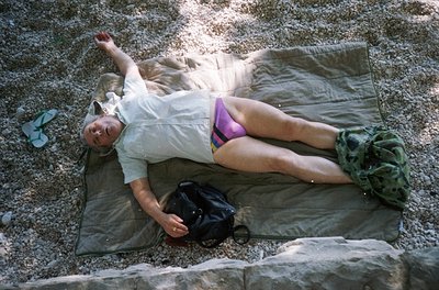 Relaxing outdoors on a green military-style tarp, individual reclines on rocky terrain with one arm raised. Light-colored shi...