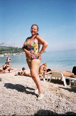 Vintage seaside scene featuring a woman in a colorful floral swimsuit posing on pebble beach. White plastic lounge chairs and...