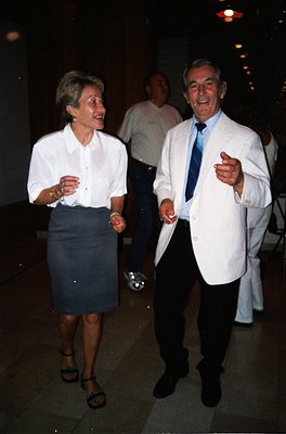 Two elegantly dressed adults laughing indoors at an evening event, likely 1990s–2000s. Woman in a white blouse, dark skirt, a...