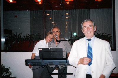 Indoor evening event featuring three performers on a stage with modernist decor. A man in a white suit and blue tie stands fr...