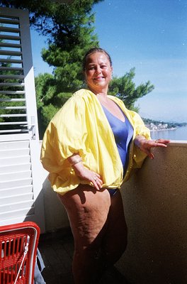Vintage seaside portrait of a woman in a bright yellow cardigan over a blue top, posing by a balcony railing. Red plastic cha...