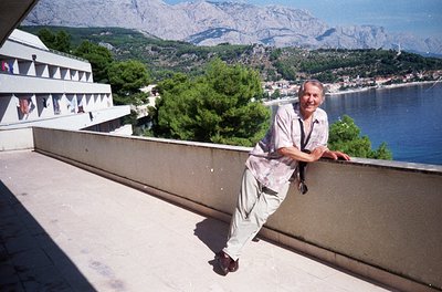 Man in retro summer attire poses on a rooftop balcony overlooking a coastal town, framed by mountains. Concrete architecture ...