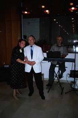 A formal evening event featuring three individuals in a well-lit indoor venue. A man in a white suit and tie stands with a wo...