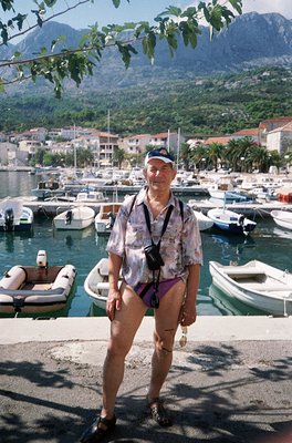 Man in 1970s-style patterned shirt and shorts poses by a Mediterranean marina, surrounded by small boats and yachts. Coastal ...