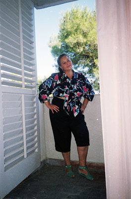 Portrait of a woman in a floral-patterned blouse with rolled sleeves, black cropped pants, and green sandals, posing in a doo...