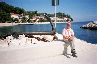 Man in light polo shirt and shorts poses beside a large rusted anchor on a seaside concrete wall, with coastal buildings and ...