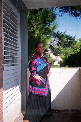 A woman stands in a shaded doorway, wearing a bold 1970s-style patterned blouse with geometric stripes in pink, blue, and bla...