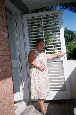 Woman in floral dress poses on concrete steps beside a white louvered door, brick wall, and lush greenery. Mid-20th century s...