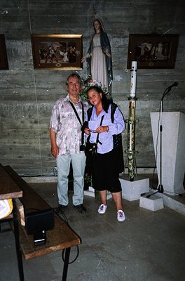 Two individuals pose indoors in a religious setting, likely a chapel or shrine. The man wears a floral-patterned shirt and li...
