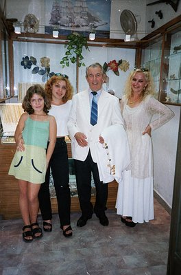 Family portrait in a vintage shop setting, likely 1980s–1990s. Four individuals pose: a man in a white suit with a striped ti...