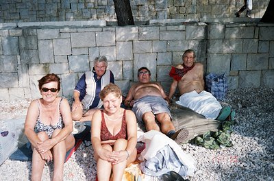 Five adults pose casually on a pebble beach beside a concrete wall, likely a seaside resort. The woman in the foreground wear...