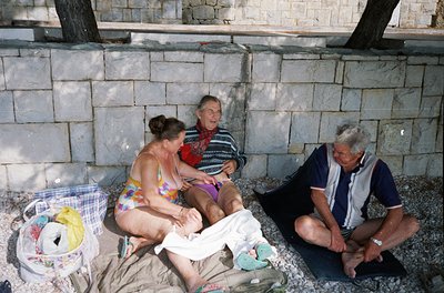 Three adults relax by a concrete wall on a pebble beach, mid-20th century. Woman in floral swimsuit kneels beside a striped-p...