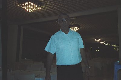 Mid-century man in a light blue button-down shirt poses indoors under a chandelier-lit ceiling. Stacked boxes and shelves sug...