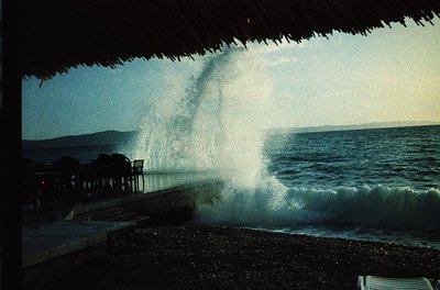 Seaside scene featuring a thatched-roof structure framing a powerful wave crashing onto a pebble beach. Ocean waves dominate ...