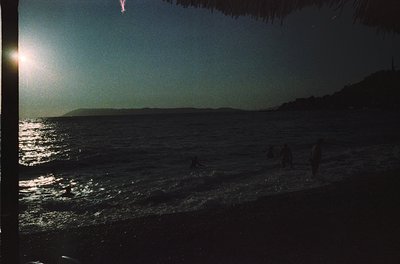 Silhouetted figures wade in shallow waves at golden-hour seaside. Low-angle sunlight reflects off water, illuminating foam. C...