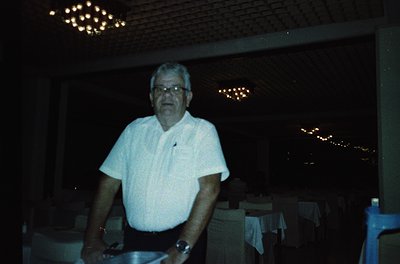Mid-century man in a white button-down shirt poses indoors, likely a restaurant or hotel lobby. Soft lighting and chandeliers...