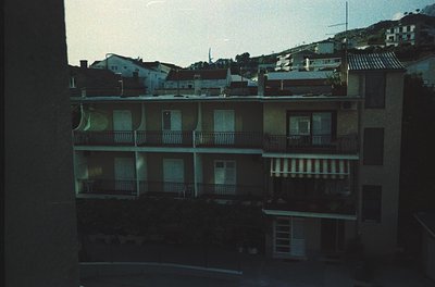 Mid-century Mediterranean urban rooftops with concrete balconies and tiled roofs, viewed from an interior window. Residential...