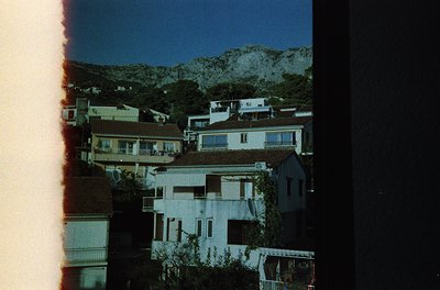 Vintage film scan of Mediterranean hillside village at dusk, featuring clustered whitewashed buildings with terracotta roofs....
