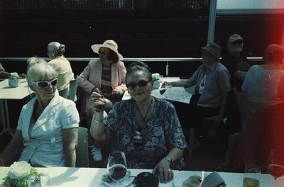 Vintage group portrait at an outdoor café, likely 1970s. Two women in center foreground wearing patterned blouses, sunglasses...
