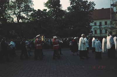 Military band in formation playing brass instruments, flanked by uniformed musicians in red jackets and white helmets. Crowd ...