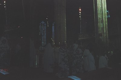 Nighttime interior of a grand, illuminated church with vertical stained-glass windows casting colored light. Ornate chandelie...