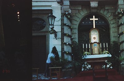 Vintage black-and-white church interior with ornate altar adorned with floral arrangements and a prominent cross. A priest in...