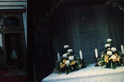 Symmetrical floral arrangements with white roses, yellow sunflowers, and greenery flank a draped altar table, lit by three li...