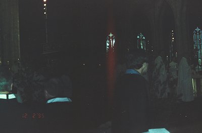 Gothic cathedral interior at night, illuminated by stained-glass windows casting colored light. Crowd seated in pews, facing ...