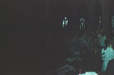 Gothic cathedral interior bathed in dim candlelight, showcasing stained-glass windows depicting religious figures. A priest i...