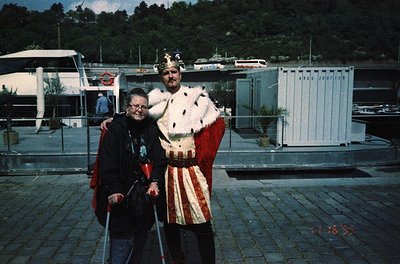 Two individuals pose outdoors near a marina, likely in the 1990s. The man wears a medieval-style fur-lined cloak and crown, s...