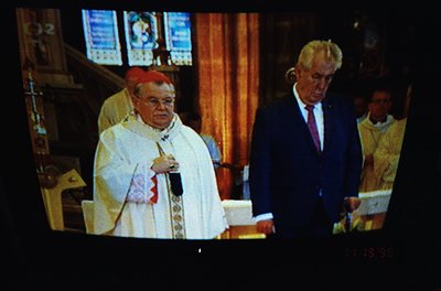 Religious ceremony featuring a cardinal in liturgical vestments conducting mass. Gothic-style church interior with stained gl...