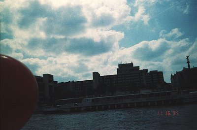 Vintage urban waterfront scene with mid-rise concrete buildings under dramatic cloud-lined sky. Bridge spans across dark wate...