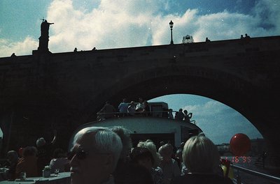 Vintage boat tour crowd on a river, likely Prague’s Vltava River, with Charles Bridge’s statues and lampposts in silhouette. ...