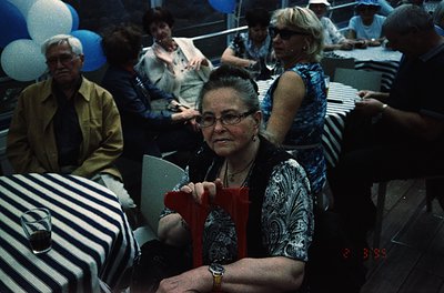 Mid-20th century social gathering in a dimly lit venue, likely a 1970s-80s event. Central figure wears patterned blouse, glas...