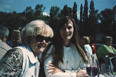 Two women share a joyful moment at an outdoor gathering, likely mid-1980s–1990s. The elder wears sunglasses and a patterned b...