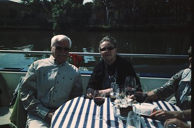 Vintage outdoor dining scene by a canal, featuring two elderly individuals seated at a striped tablecloth. Glasses of red win...