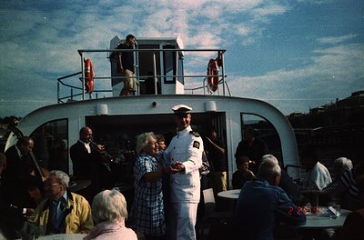 Mid-20th century ferry deck scene with uniformed captain greeting passengers. White vessel features lifebuoys on railings, ur...