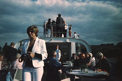 Vintage yacht deck party with mid-century attire. A woman in a white blazer holds a clipboard, overseeing guests seated at ro...