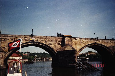 Historic stone bridge with Gothic arches spanning a river, featuring a flag with a red/blue emblem. Pedestrians and a boat wi...