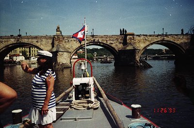 Vintage boat operator in striped uniform waves from Prague’s Charles Bridge, 1980s-90s. Gothic arches, historic flag, and riv...
