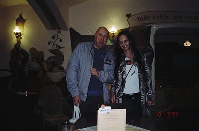 Two individuals pose indoors beside a vintage car, likely a Škoda, in a dimly lit setting with rustic decor. The man wears a ...