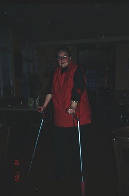 Vintage black-and-white photo of elderly individual in a red vest, holding cane-like sticks, indoors near kitchen appliances....
