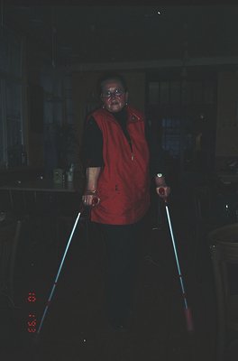 Indoor portrait of an elderly woman using illuminated walking sticks in low-light, likely a 1970s–1980s kitchen. She wears a ...