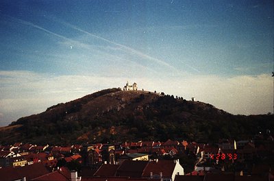 Vintage aerial view of a hilltop fortress atop a forested hill, surrounded by a densely built European town. The structure fe...