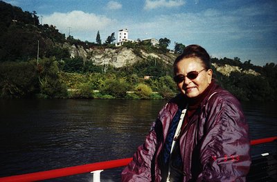 Woman in maroon jacket poses by a riverbank, wearing sunglasses and a lanyard. Rocky hillside with sparse vegetation and a wh...
