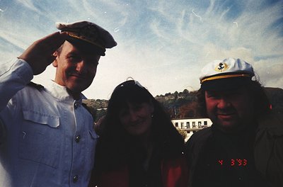 Three individuals pose outdoors in vintage attire, likely 1990s. Left: man in white naval-style cap and jacket. Center: woman...